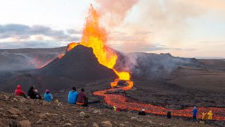 Um grupo de pessoas observa de longe um vulcão explodir com lava