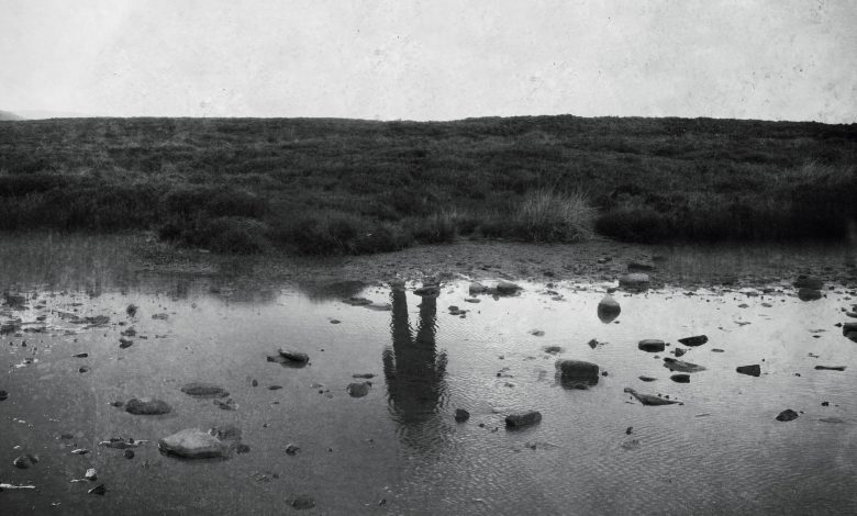 Uma foto em preto e branco de uma margem vazia de um rio, com a figura de um homem refletida na água