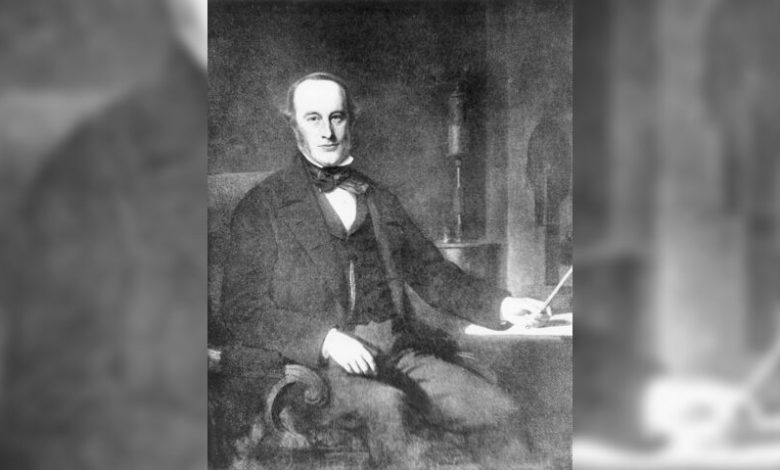 A black-and-white image of a white man sitting on a chair in a tuxedo